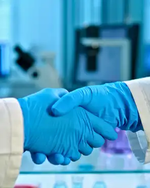 Two scientists in lab coats and blue gloves shake hands in a lab. Background includes scientific equipment like monitors and glassware, indicating collaboration in medical research.