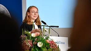 Zeynep Akata steht an einem Rednerpult der Leopoldina und spricht in ein Mikrophon. Hinter ihr steht eine blaue Messewand mit Leopoldina-Logo.